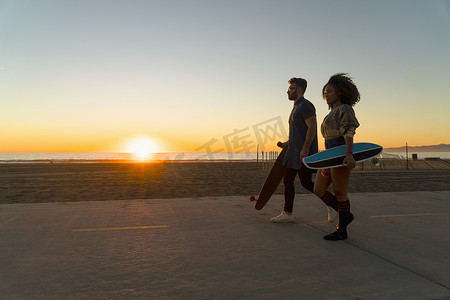 一对情侣沿着海滩边的小路走着手里拿着滑板