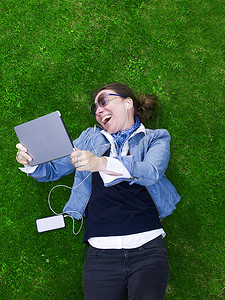 俯瞰成年女子手持数字平板电脑躺在草地上笑着