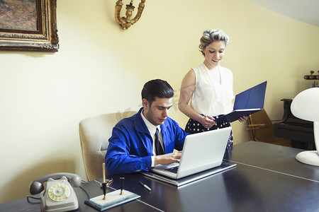 复古角摄影照片_年轻的复古夫妇坐在办公桌前拿着文件和笔记本电脑