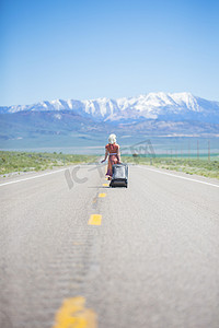 美国内华达州50年代风格的年轻女子拖着轮式行李箱独自走在50号高速公路上的后景