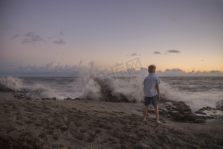 日出时男孩看着溅起的海浪的后景美国佛罗里达州朱庇特岛吹石保护区