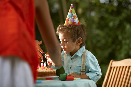 可爱生日蛋糕摄影照片_母亲和儿子在花园生日聚会上吹生日蛋糕上的蜡烛