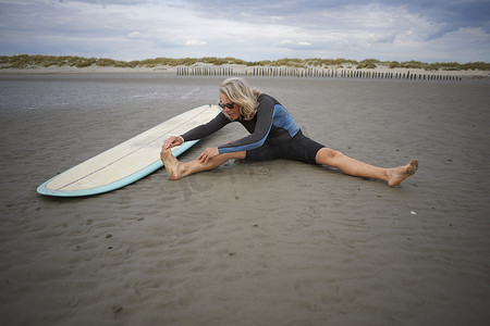 老年人防护摄影照片_老妇人坐在沙滩上伸展四肢身旁有冲浪板