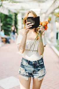 一名年轻女子站在户外手持玫瑰使用智能手机