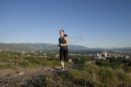 公路奔跑摄影照片_年轻的女跑步者沿着山谷中城市上空的跑道奔跑