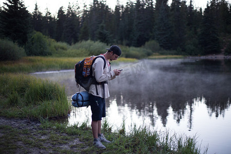 森林摄影照片_男性徒步旅行者在美国俄勒冈州胡德山国家森林迷雾湖边阅读智能手机短信