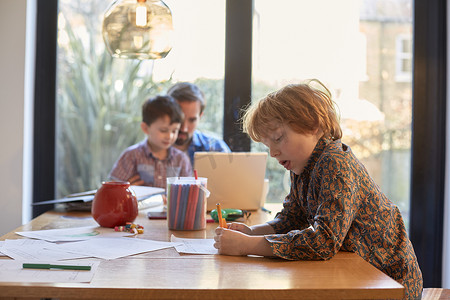 男孩在餐桌上涂颜色而父亲和兄弟一起使用笔记本电脑