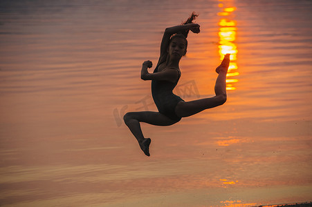 夕阳下海边身影的女孩在半空中跳跃双腿分开看着摄像机