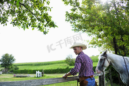 带着牛仔帽摄影照片_一名年轻男子穿着牛仔装备带着马检查栅栏