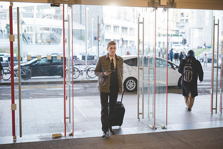 商务行走摄影照片_提着手提箱穿过玻璃门的年轻商人