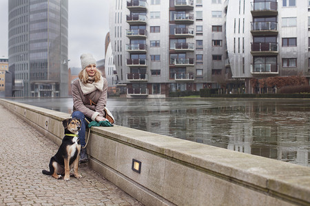 坐在河边摄影照片_中年妇女和她的狗坐在河边的墙上
