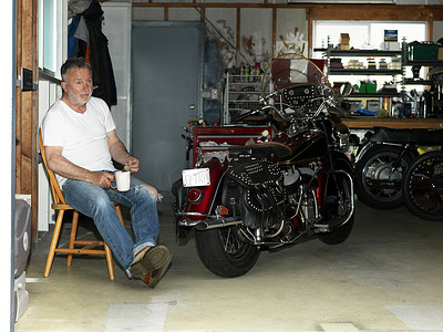 骑摩托车赛车摄影照片_一位年长的男子坐在车库里手里拿着咖啡骑着摩托车