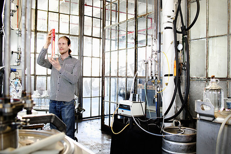 酿酒车间摄影照片_年轻男性伏特加酿酒工人在酿酒车间测量钢瓶中的液体