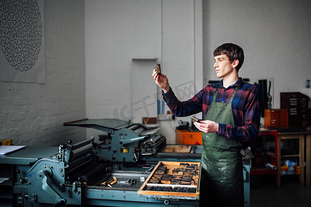 印刷机摄影照片_年轻的工匠在印刷机旁在印刷车间里举起信件