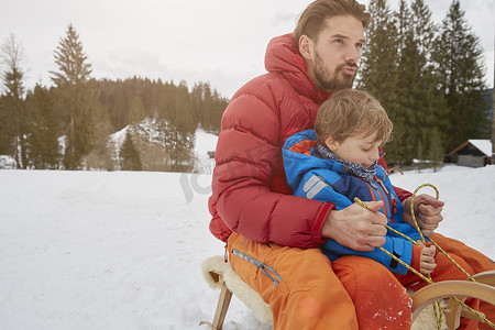 保暖衣物摄影照片_德国巴伐利亚州埃尔茂市一对年轻男子和儿子在雪地里滑雪橇