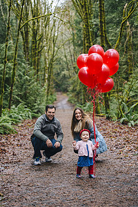 科技2.5装饰摄影照片_父母在森林里蹲着蹒跚学步的女儿手持一堆红色气球的照片