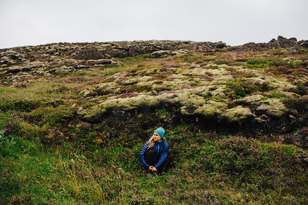 冰岛一名中年妇女戴着针织帽蹲在土丘前目光投向别处