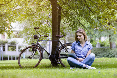 使用数字平板电脑的年轻女子盘腿坐在自行车旁边的草地上