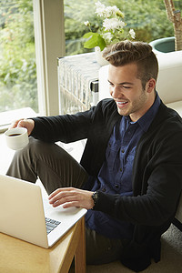 微笑的男人坐在家里的地板上看着笔记本电脑喝着一杯咖啡