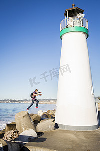 极限滑板摄影照片_灯塔旁一名年轻人手持滑板从防波堤上跳下