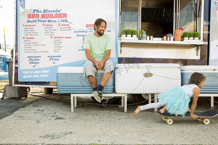 父亲坐在快餐拖车旁看女儿玩滑板