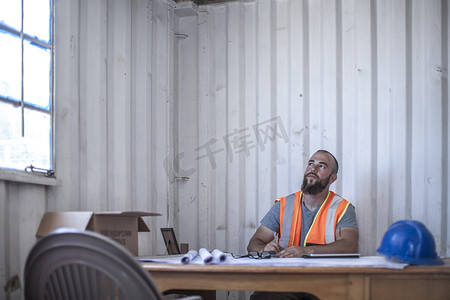 职场沟通技巧摄影照片_建筑工人坐在轻便小屋的办公桌前