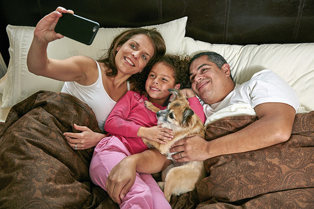 高角看女孩与父母和狗在床上用智能手机自拍微笑