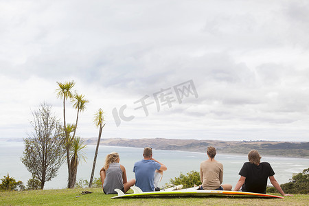 四名年轻的成年冲浪者坐在草地上望向大海