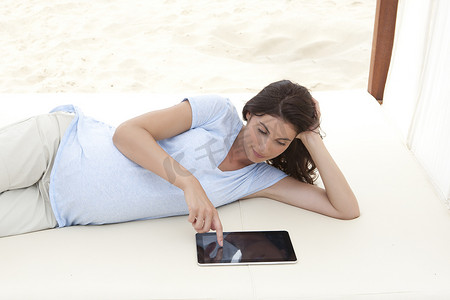 使用数字平板电脑躺在沙滩上的怀孕中年妇女