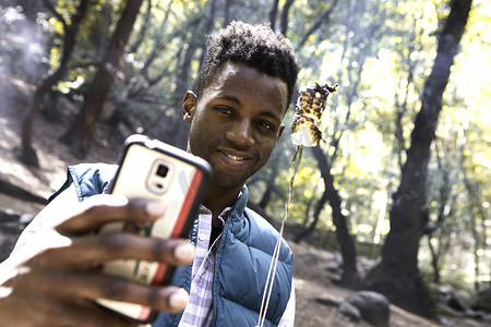 吉利特摄影照片_一名年轻的男性徒步旅行者在美国加利福尼亚州阿卡迪亚的森林里与烤棉花糖智能手机自拍