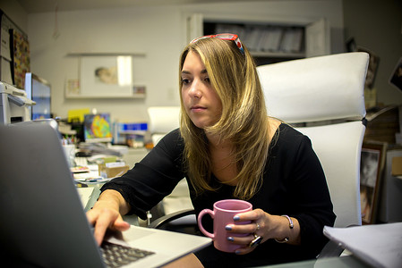 办公室里的年轻女性坐在办公桌前拿着咖啡杯使用笔记本电脑
