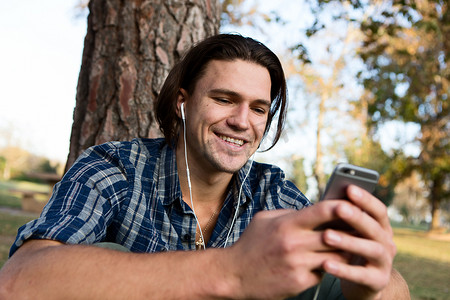 一位戴着耳机的年轻人坐在树旁微笑着看着智能手机