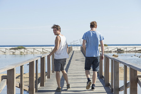两个人抬头看摄影照片_意大利撒丁岛年轻人走在木制码头上抬头看着肩膀的全景
