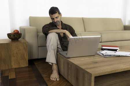 赤脚的成熟男子拿着笔记本电脑坐在木地板上俯视着