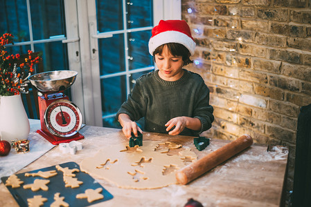 戴圣诞帽的男孩摄影照片_戴圣诞帽的男孩在厨房柜台准备圣诞饼干