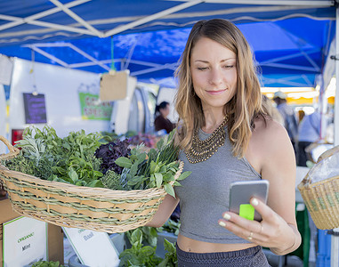 一名女子在水果和蔬菜摊上拿着一篮子新鲜的草药看着智能手机