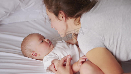 可爱的年轻妈妈抱着甜美可爱的女婴躺在床上笑容可亲的母亲和可爱的小女孩抱在卧室里母亲和孩子的柔情时刻
