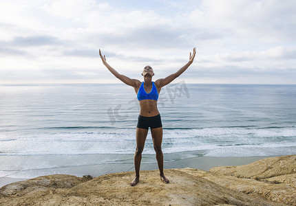 举起武器摄影照片_站在海边岩石上的年轻女子的肖像举起双臂抬头看