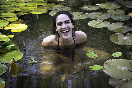 一名年轻女子坐在池塘里面带微笑地看着镜头身旁围着百合花