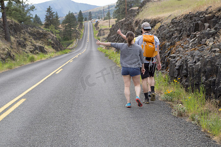 高尔夫球包摄影照片_一对年轻夫妇沿着公路徒步旅行试图抬起拇指后视