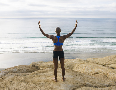 年轻女子站在海边的岩石上举起双臂向后看
