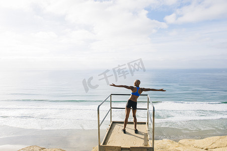 年轻女子站在海边的瞭望点上举起双臂向后看