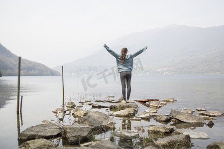举起武器摄影照片_意大利皮埃蒙特的湖上举起双臂的年轻女子在岩石上的背影