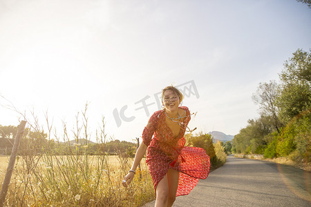 公路奔跑摄影照片_快乐的年轻女子穿着红色连衣裙在乡村公路上奔跑西班牙马略卡岛