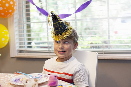 戴着派对帽的男孩坐在桌子边吃生日蛋糕拉着一张鬼脸