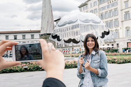 拍照旅游景点摄影照片_意大利托斯卡纳佛罗伦萨圣玛丽亚·诺维拉广场一名撑着伞的年轻女子用智能手机摆姿势给朋友拍照