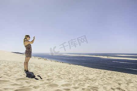 迈特凯摄影照片_用智能手机拍摄大海的年轻女子法国皮拉特沙丘