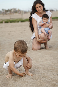 儿童培育摄影照片_蹒跚学步的孩子用手在沙子里挖掘背景是母亲和婴儿