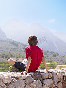 山水风景图案摄影照片_坐在石墙上凝视风景的男孩西班牙马略卡岛