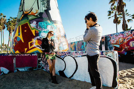 年轻女子站在海滩上的涂鸦艺术旁年轻男子给她拍照